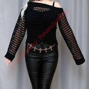 Op de afbeelding: Zwarte, open geweven, off-the-shoulder top met lange mouwen. De top heeft een uniek uitgesneden ontwerp en wordt gecombineerd met een zwarte broek en een zilveren kruisjeskettingriem. De algehele stijl is goth.