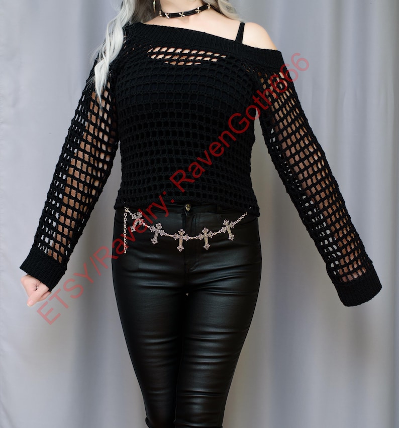 Op de afbeelding: Zwarte, open geweven, off-the-shoulder top met lange mouwen. De top heeft een uniek uitgesneden ontwerp en wordt gecombineerd met een zwarte broek en een zilveren kruisjeskettingriem. De algehele stijl is goth.