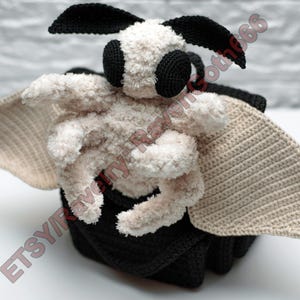 May include: A handcrafted crochet moth toy. The moth features a soft, cream-coloured body, large beige wings, and black details on the eyes and antennae. It rests on a black base. The Etsy shop name is visible in the image.