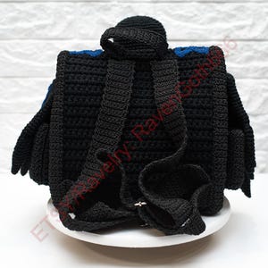 May include: Black crochet backpack with blue accents. The backpack features adjustable straps and a top handle. The design includes a flap and side pockets. The bag is displayed on a white surface against a white brick wall.