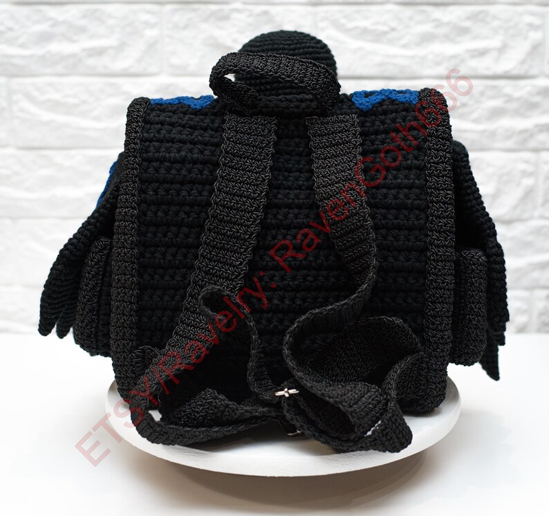 May include: Black crochet backpack with blue accents. The backpack features adjustable straps and a top handle. The design includes a flap and side pockets. The bag is displayed on a white surface against a white brick wall.
