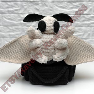 May include: A handmade crochet moth plushie. It has a black body and head, cream-colored wings, and a fluffy cream-colored body. The moth has large black eyes and antennae. The wings are a light tan colour. The plushie is on a white surface.