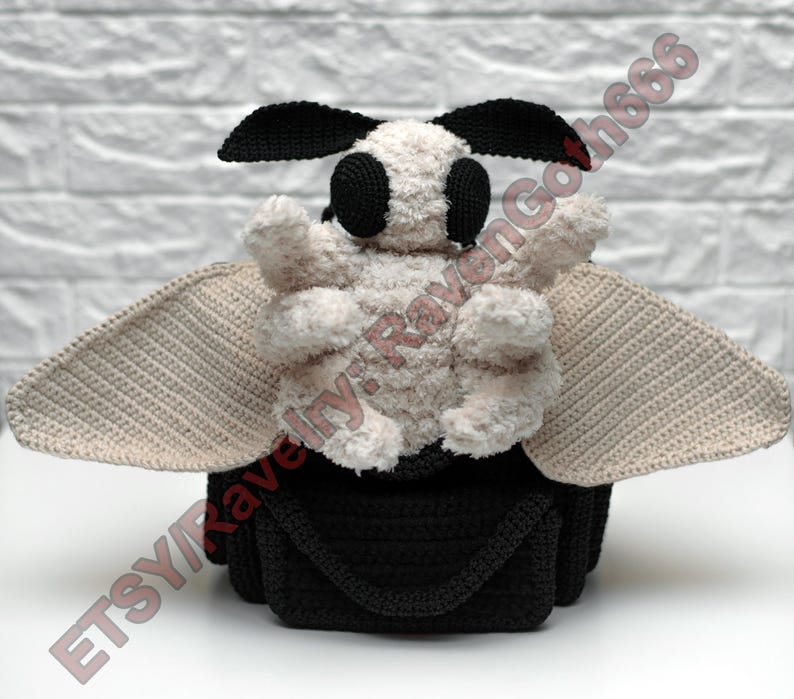 May include: A handmade crochet moth plushie. It has a black body and head, cream-colored wings, and a fluffy cream-colored body. The moth has large black eyes and antennae. The wings are a light tan colour. The plushie is on a white surface.