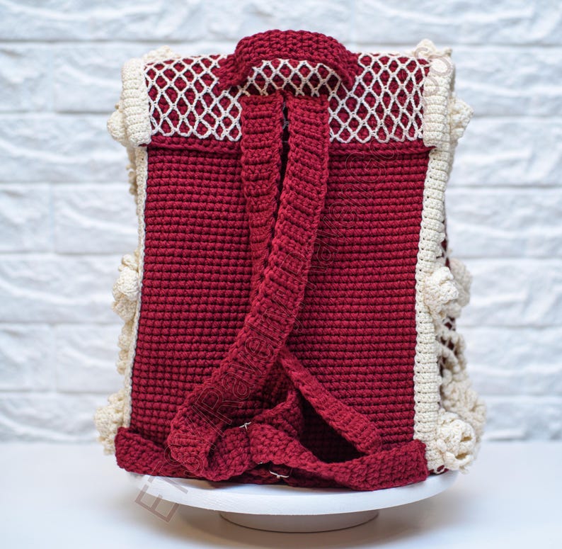 K&ouml;nnte beinhalten: Ein handgefertigter H&auml;kelrucksack in tiefroter Farbe mit cremefarbenen Verzierungen und einem wei&szlig;en Netzdesign auf der oberen Klappe. Der Rucksack hat verstellbare Tr&auml;ger und einen Griff.