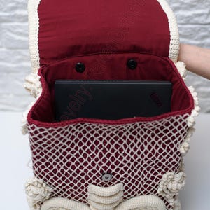 K&ouml;nnte beinhalten: Handgefertigter H&auml;kelrucksack in Burgunderrot und Creme. Die Tasche hat ein gitterartiges Muster mit cremefarbenen Verzierungen und dekorativen Akzenten. Das Innere ist mit burgunderfarbenem Stoff gef&uuml;ttert, und ein schwarzer Laptop ist sichtbar.