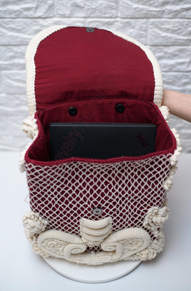 K&ouml;nnte beinhalten: Handgefertigter H&auml;kelrucksack in Burgunderrot und Creme. Die Tasche hat ein gitterartiges Muster mit cremefarbenen Verzierungen und dekorativen Akzenten. Das Innere ist mit burgunderfarbenem Stoff gef&uuml;ttert, und ein schwarzer Laptop ist sichtbar.