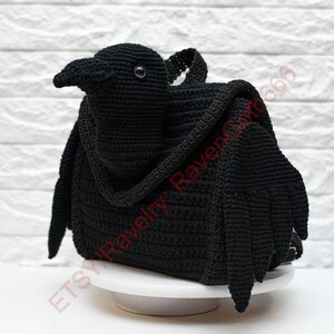 May include: A black crochet raven-shaped bag with a handle and wings. The bag is sitting on a white cake stand against a white brick wall. The Etsy shop name "Etsy Ravelry Raven666" is visible.