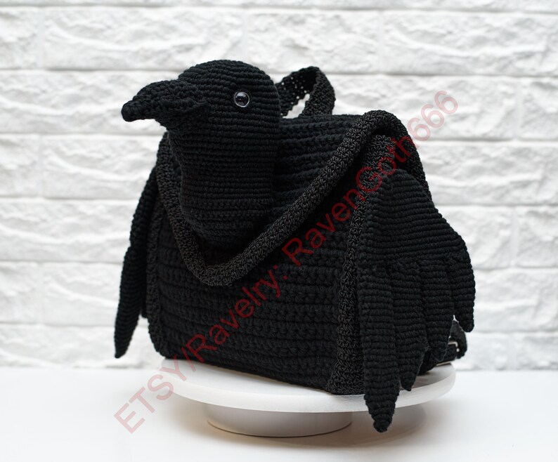May include: A black crochet raven-shaped bag with a handle and wings. The bag is sitting on a white cake stand against a white brick wall. The Etsy shop name "Etsy Ravelry Raven666" is visible.