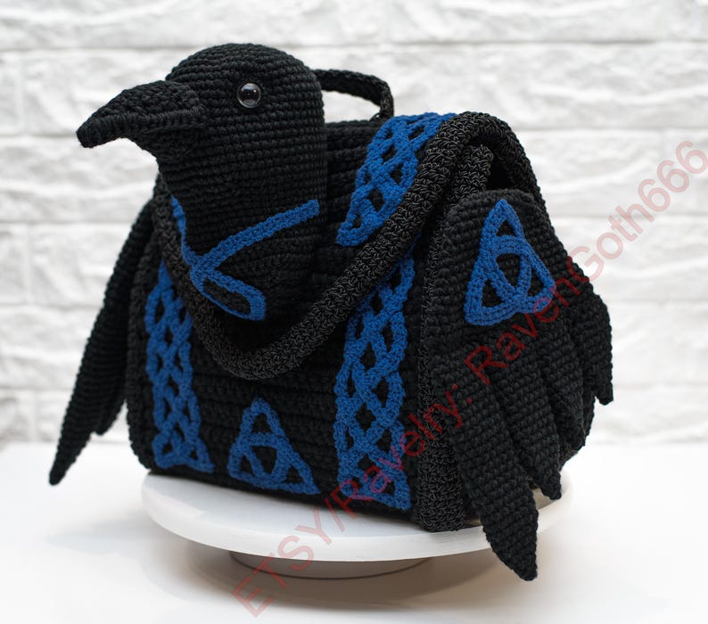May include: A black crochet raven-shaped bag with blue Celtic knot accents. The bag features a handle and wings, and the design incorporates blue trim and details. The raven's head is detailed with an eye and beak.