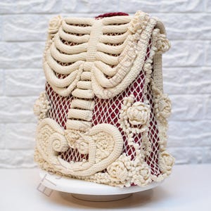 K&ouml;nnte beinhalten: Eine cremefarbene H&auml;kelh&uuml;lle mit einem Brustkorb-Design auf der Vorderseite. Sie hat ein bordeauxfarbenes Netzinnenfutter und ist mit floralen und scrollartigen H&auml;keldetails verziert. Der Artikel wird auf einem wei&szlig;en Sockel pr&auml;sentiert.