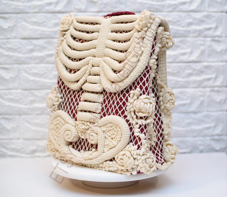 K&ouml;nnte beinhalten: Eine cremefarbene H&auml;kelh&uuml;lle mit einem Brustkorb-Design auf der Vorderseite. Sie hat ein bordeauxfarbenes Netzinnenfutter und ist mit floralen und scrollartigen H&auml;keldetails verziert. Der Artikel wird auf einem wei&szlig;en Sockel pr&auml;sentiert.