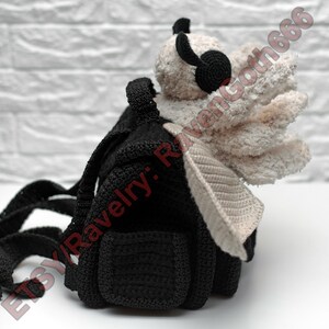 May include: A black crochet backpack with a cream-coloured wing and a small, black, plush head with black headphones. The backpack has adjustable straps and a small pocket. The Etsy shop name "Etsy: RavenGoth666" is visible.