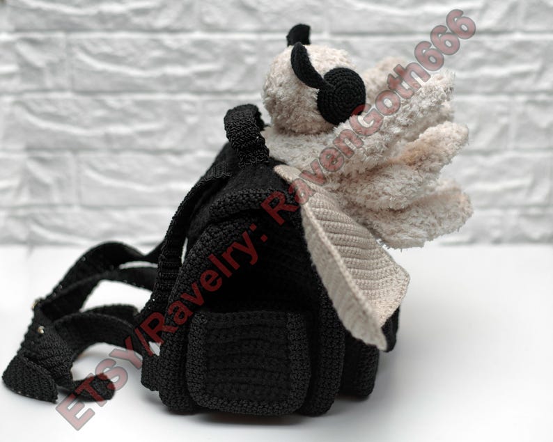 May include: A black crochet backpack with a cream-coloured wing and a small, black, plush head with black headphones. The backpack has adjustable straps and a small pocket. The Etsy shop name "Etsy: RavenGoth666" is visible.