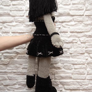May include: A gray crocheted doll with black yarn hair, wearing a black dress with white stitching and black boots. The doll has a cat-like face and ears.