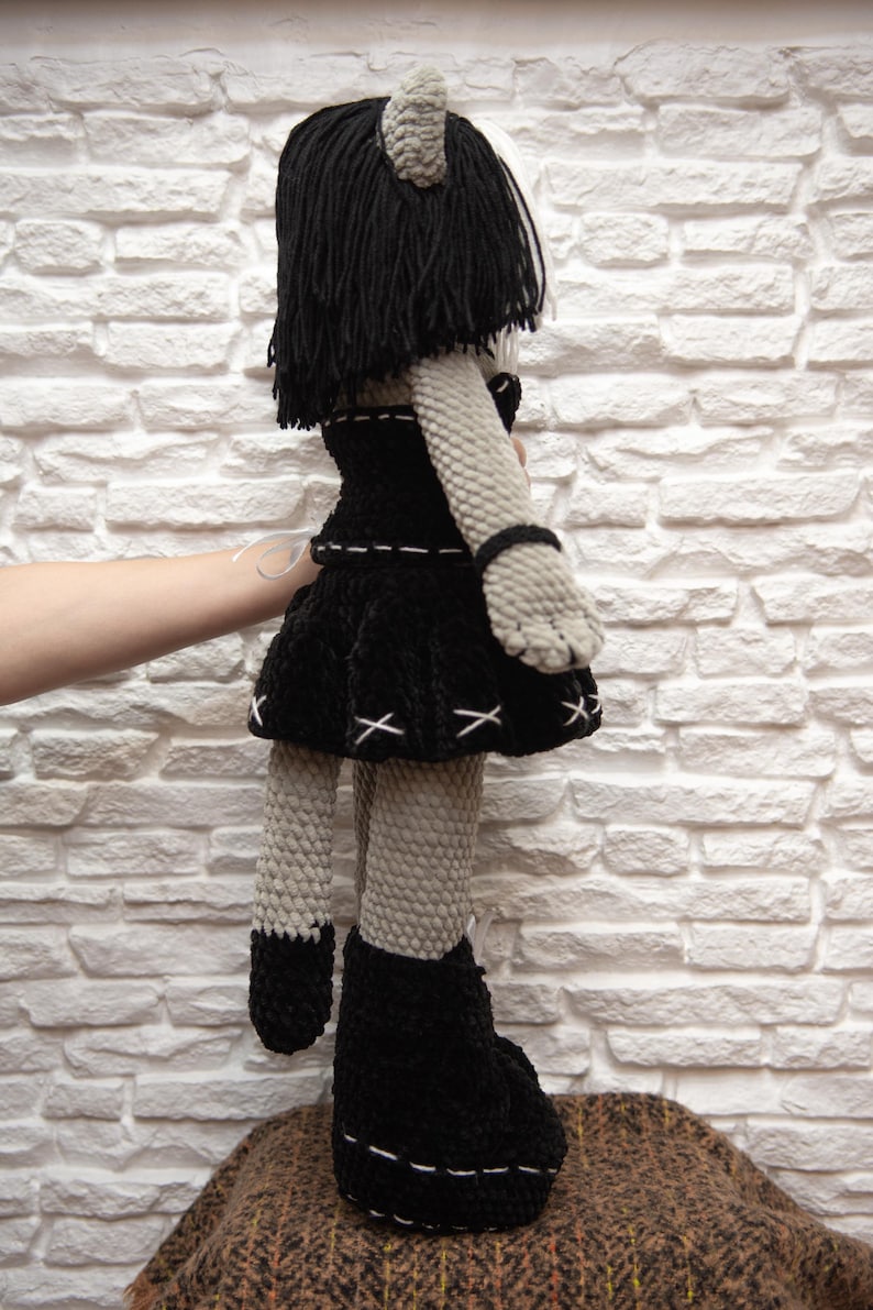 May include: A gray crocheted doll with black yarn hair, wearing a black dress with white stitching and black boots. The doll has a cat-like face and ears.
