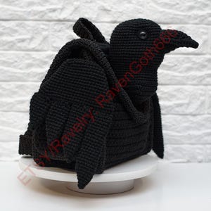 May include: A black crochet raven-shaped backpack with wings and a beak. The handmade bag is sitting on a white surface against a white brick wall. The raven has a detailed, textured appearance.