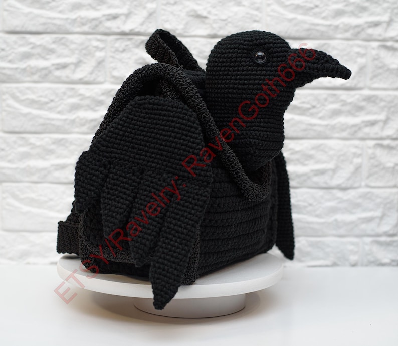 May include: A black crochet raven-shaped backpack with wings and a beak. The handmade bag is sitting on a white surface against a white brick wall. The raven has a detailed, textured appearance.