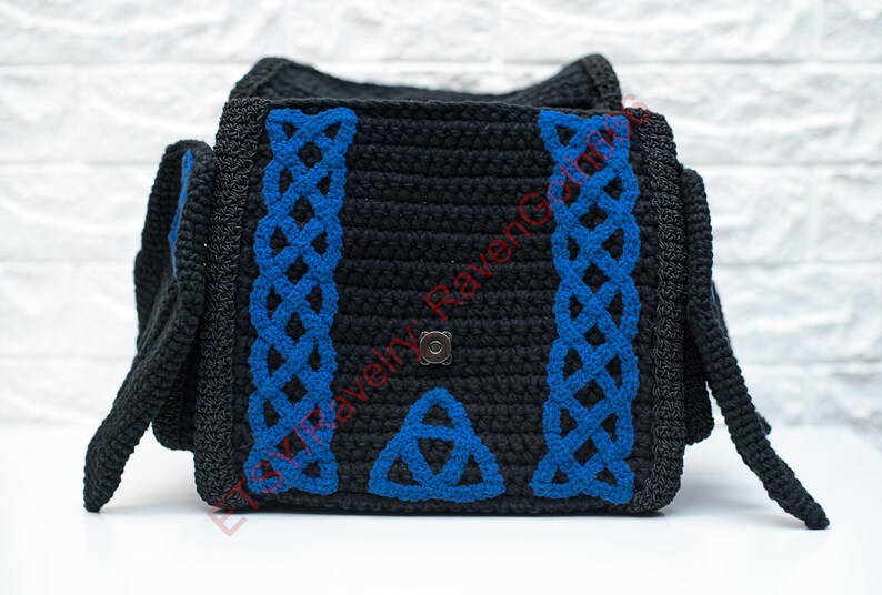 May include: Black crochet bag with blue Celtic knotwork design. The bag features a flap closure and side panels. The blue knotwork is on the front and sides of the bag. The bag is sitting on a white surface.