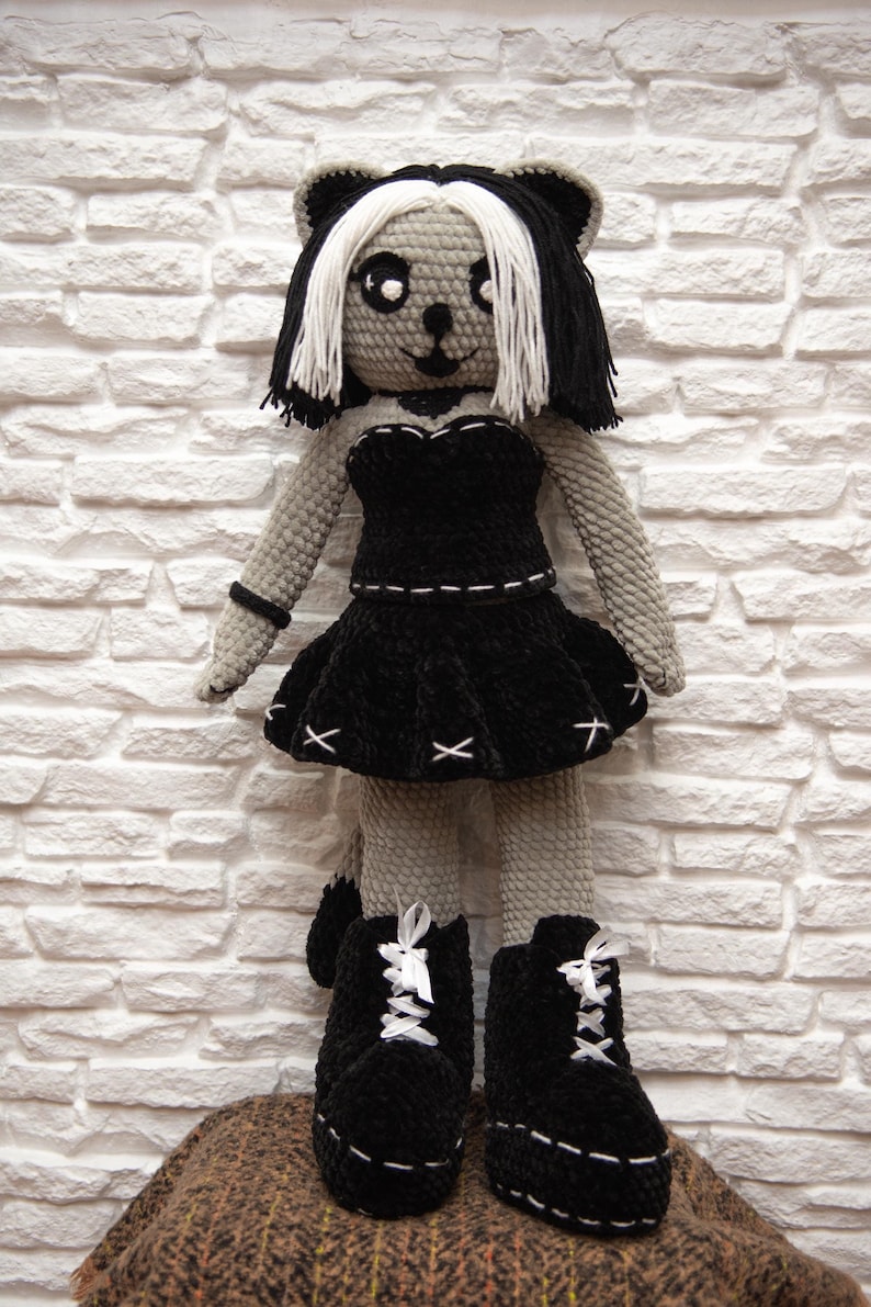 May include: A crocheted plush toy with black and white hair, wearing a black dress and black boots. The toy is standing on a brown surface.