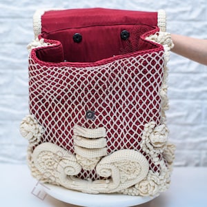 K&ouml;nnte beinhalten: Handgefertigte H&auml;keltasche mit bordeauxrotem Innenraum und cremefarbenem, durchbrochenem &Auml;u&szlig;eren. Die Tasche hat dekorative cremefarbene Blumen- und Schn&ouml;rkelakzente. Die Tasche ist offen und zeigt das Innere, und wird mit einem Magnetverschluss geschlossen.