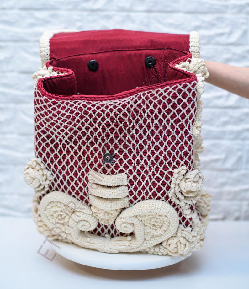 K&ouml;nnte beinhalten: Handgefertigte H&auml;keltasche mit bordeauxrotem Innenraum und cremefarbenem, durchbrochenem &Auml;u&szlig;eren. Die Tasche hat dekorative cremefarbene Blumen- und Schn&ouml;rkelakzente. Die Tasche ist offen und zeigt das Innere, und wird mit einem Magnetverschluss geschlossen.