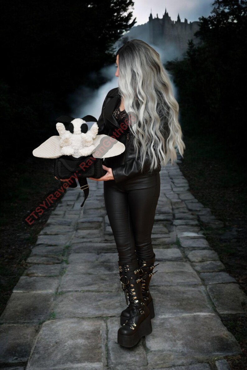 May include: A black and white moth-shaped plush toy with large wings is held in front of a dark background. The plush toy is the focal point, with a person wearing black leather clothing and platform boots in the background. The image has a gothic aesthetic.