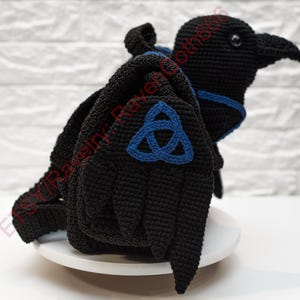 May include: A black crochet raven with blue accents and a blue Celtic knot design on the wings. The raven is perched on a white surface against a white brick background. The raven's wings are spread, and it has a detailed beak.