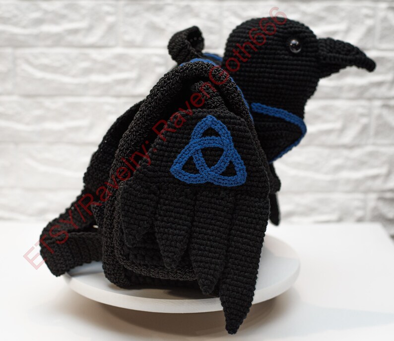 May include: A black crochet raven with blue accents and a blue Celtic knot design on the wings. The raven is perched on a white surface against a white brick background. The raven's wings are spread, and it has a detailed beak.