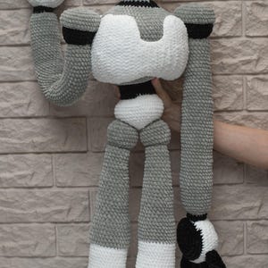May include: A crocheted robot toy with a gray and white body, black arms and legs, and a black head. The robot is standing on a white surface.