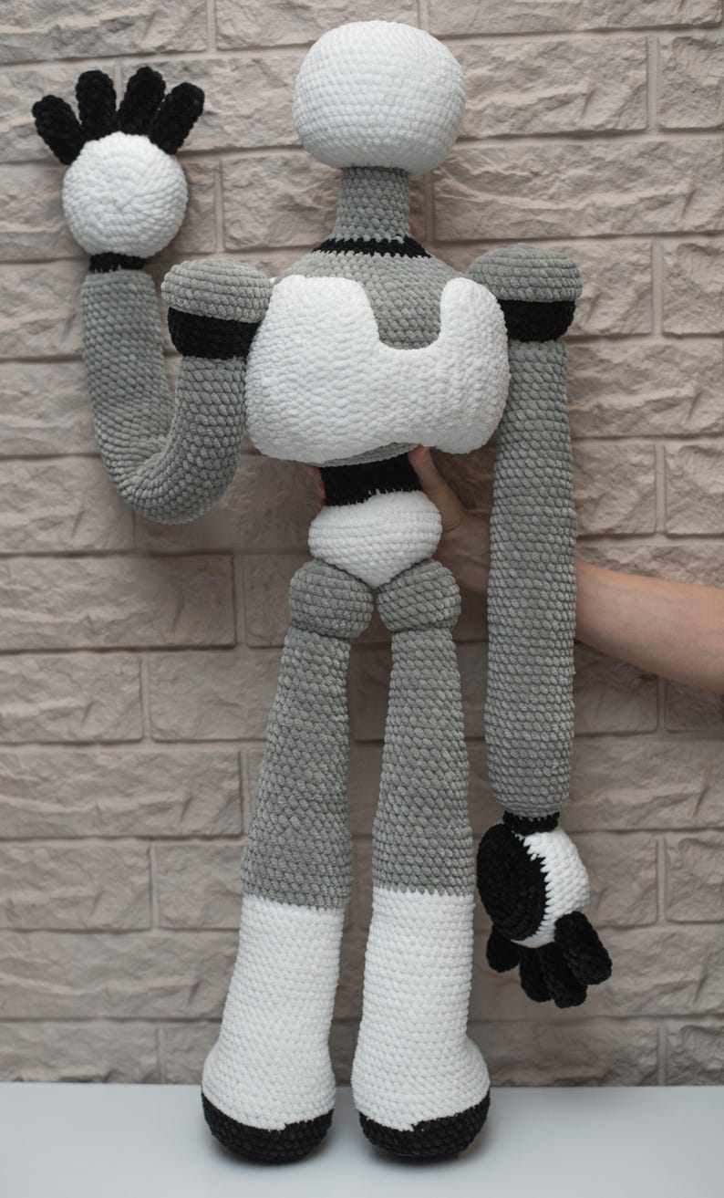 May include: A crocheted robot toy with a gray and white body, black arms and legs, and a black head. The robot is standing on a white surface.