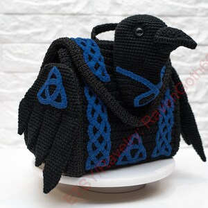 May include: A black crochet bag shaped like a raven, with blue Celtic knot accents. The bag features a handle and wings, and is displayed on a white surface. The raven's head is detailed with a beak and eye.