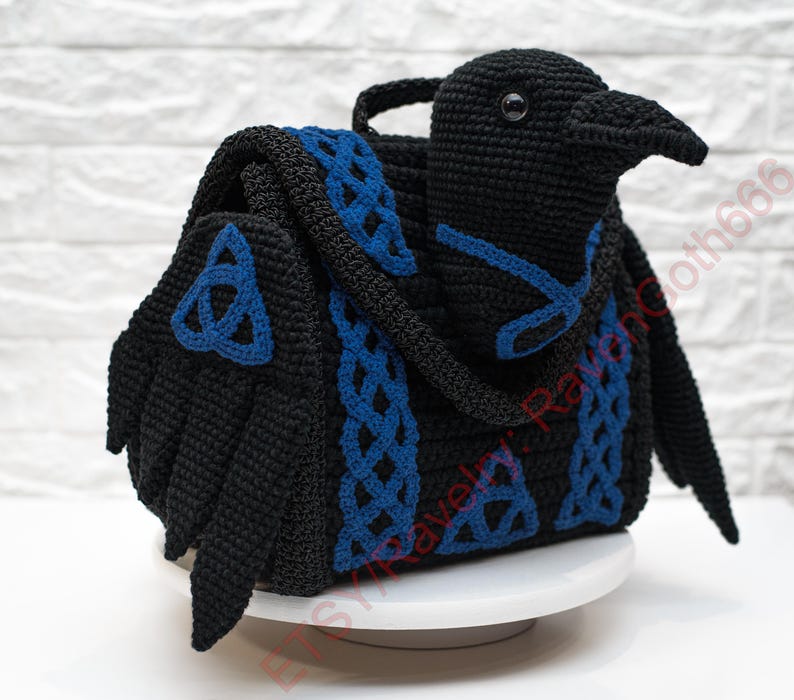 May include: A black crochet bag shaped like a raven, with blue Celtic knot accents. The bag features a handle and wings, and is displayed on a white surface. The raven's head is detailed with a beak and eye.