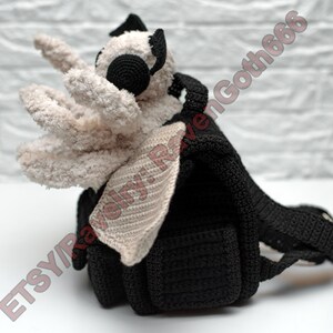 May include: A handmade black crochet backpack with a plush, cream-coloured creature attached. The creature has large, round black eyes and a pair of cream-coloured, crocheted wings. The backpack has adjustable straps and a small pocket.