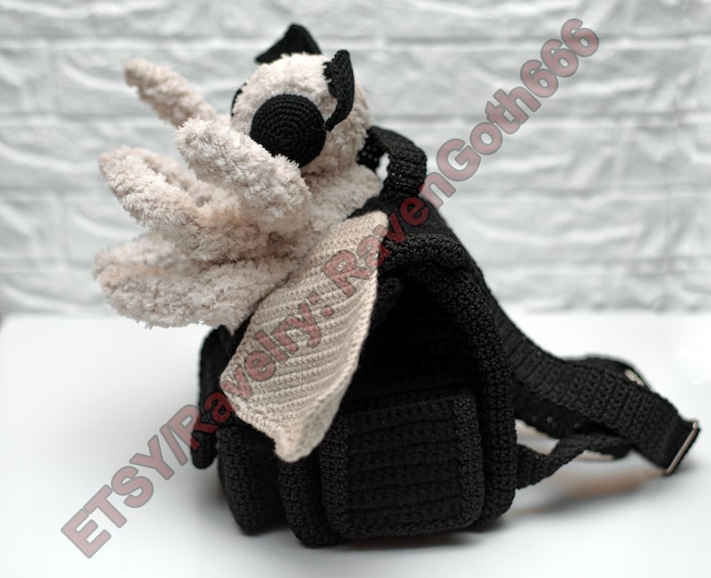 May include: A handmade black crochet backpack with a plush, cream-coloured creature attached. The creature has large, round black eyes and a pair of cream-coloured, crocheted wings. The backpack has adjustable straps and a small pocket.