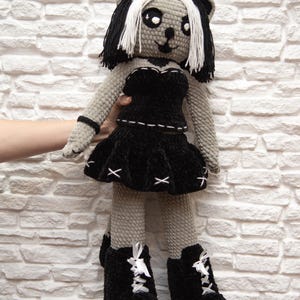 May include: A gray crocheted doll with black and white hair wearing a black dress and black boots. The doll is standing on a brown and black patterned surface.