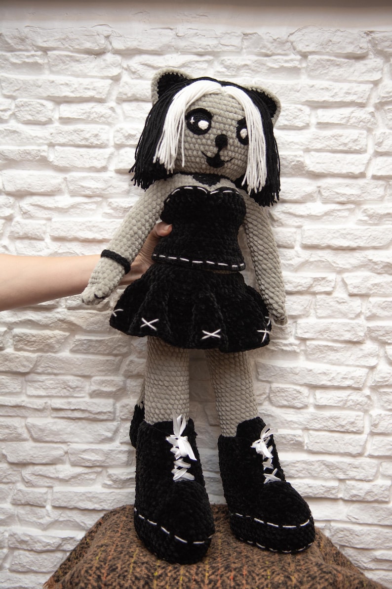 May include: A gray crocheted doll with black and white hair wearing a black dress and black boots. The doll is standing on a brown and black patterned surface.