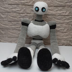 May include: A crocheted robot toy with a white body, gray arms and legs, and black feet. The robot has a white head with two blue eyes.