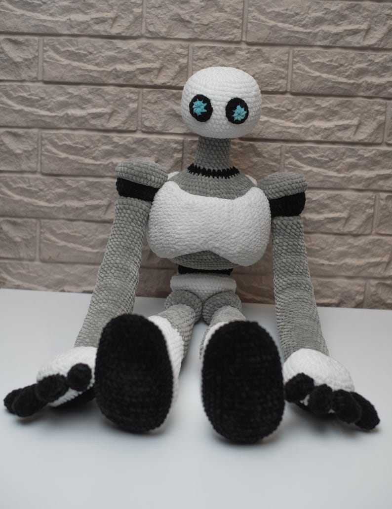 May include: A crocheted robot toy with a white body, gray arms and legs, and black feet. The robot has a white head with two blue eyes.
