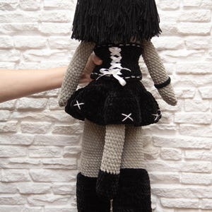 May include: A gray crocheted doll with black hair, wearing a black dress with a white lace-up corset and black boots. The doll is standing on a brown and black patterned surface.