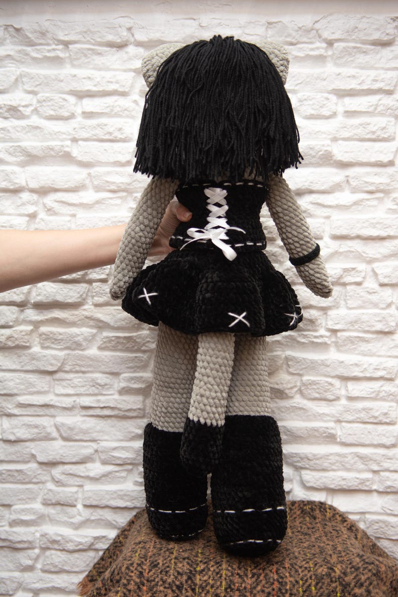 May include: A gray crocheted doll with black hair, wearing a black dress with a white lace-up corset and black boots. The doll is standing on a brown and black patterned surface.