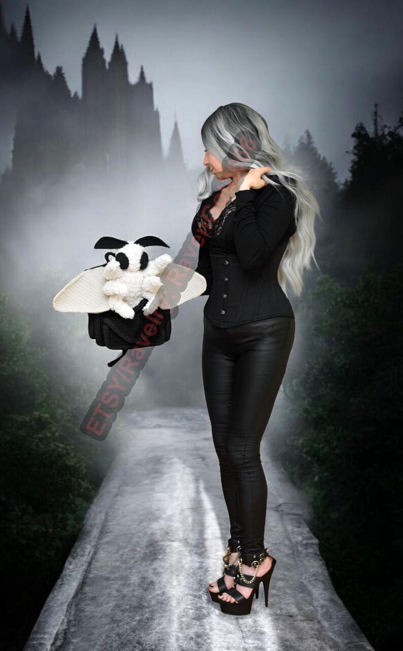 May include: A crochet moth plushie in black and white, with large wings. The moth is held by a person in a black outfit. The background shows a misty forest and a castle silhouette.