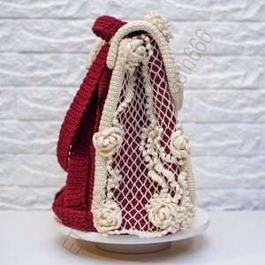 K&ouml;nnte beinhalten: Handgefertigte H&auml;keltasche in Burgunderrot und Creme. Die Tasche hat ein netzartiges Design mit cremefarbenen Blumenverzierungen. Der Riemen und der Hauptteil der Tasche sind burgunderrot. Die Tasche wird auf einer wei&szlig;en Oberfl&auml;che pr&auml;sentiert.