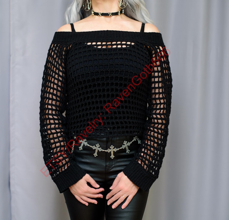 Op de afbeelding: Zwarte off-the-shoulder gehaakte trui met lange mouwen. De trui heeft een mesh-ontwerp en wordt gecombineerd met een zwarte leren broek en een zilveren kruiskettingriem. De top wordt over een zwarte camisole gedragen.