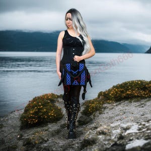 May include: A woman in a black tank top and corset, with a blue and red patterned skirt, and black boots. The background features a lake and mountains under a cloudy sky. The outfit has a gothic or fantasy theme.