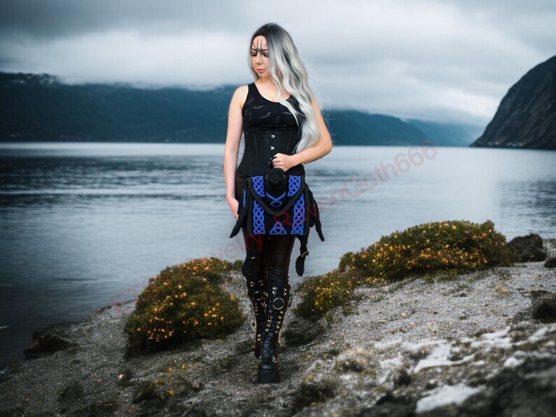 May include: A woman in a black tank top and corset, with a blue and red patterned skirt, and black boots. The background features a lake and mountains under a cloudy sky. The outfit has a gothic or fantasy theme.