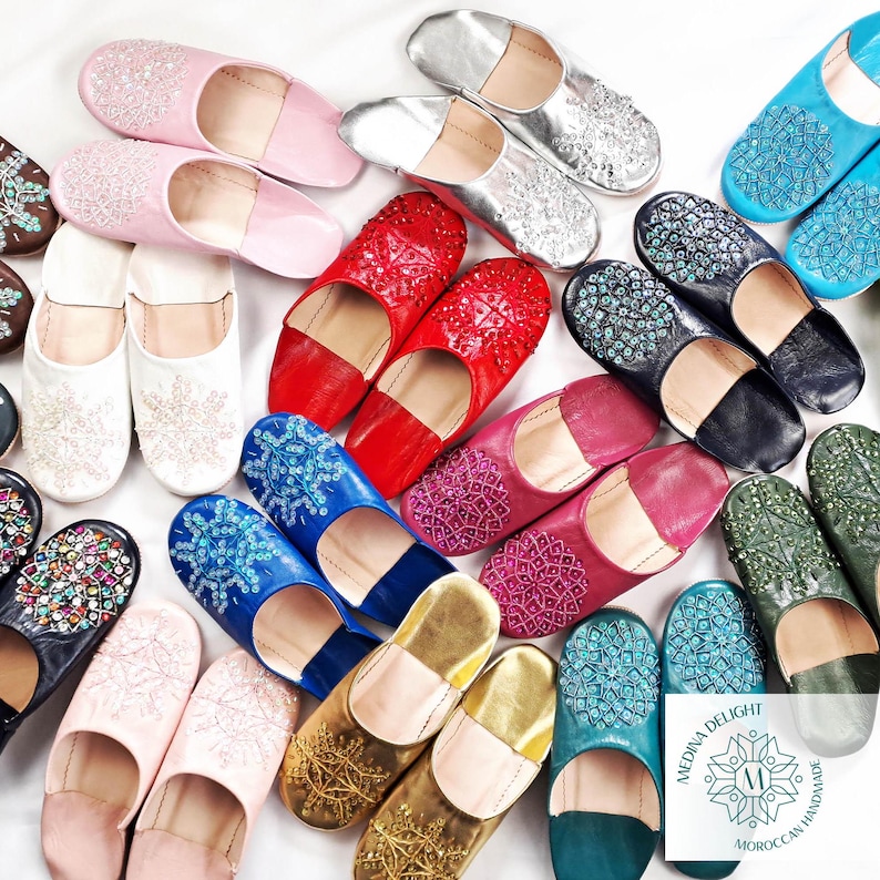May include: Assortment of colorful Moroccan leather slippers with intricate beaded designs. The slippers are handmade and feature a variety of colors, including pink, blue, green, gold, and red.
