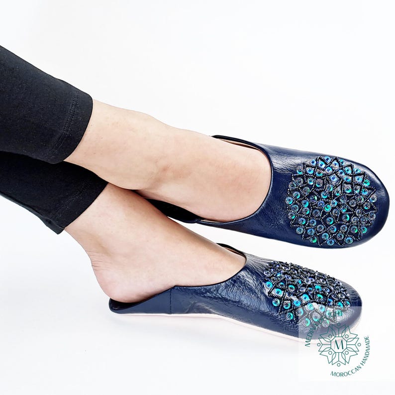 May include: Pair of navy blue leather slippers with a decorative pattern of turquoise and green sequins. The slippers have a pointed toe and a flat sole. The brand name "MOROCCAN HANDMADE" is printed on the sole.