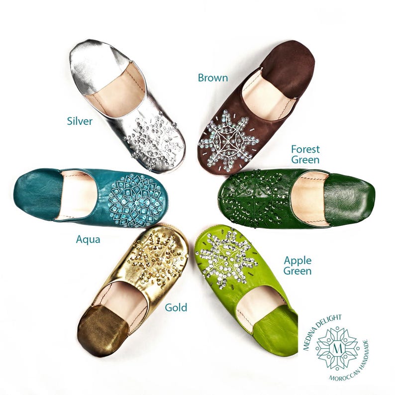 May include: Six pairs of leather slippers with jeweled embellishments in various colors, including silver, aqua, gold, forest green, apple green, and brown. The slippers are arranged in a circle on a white background. The text "Medina Delight Moroccan Handmade" is visible in the lower right corner.