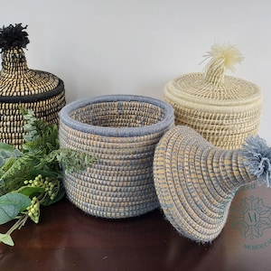 Op de afbeelding: Drie geweven opbergmanden met deksels en pompoenen. De manden zijn gemaakt van natuurlijke materialen en hebben verschillende kleuren, waaronder zwart, wit en blauw.