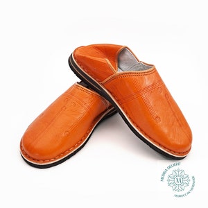 Puede incluir: Un par de zapatillas de cuero naranja con ribete negro. Las zapatillas tienen un diseño acolchado y están hechas en Marruecos.